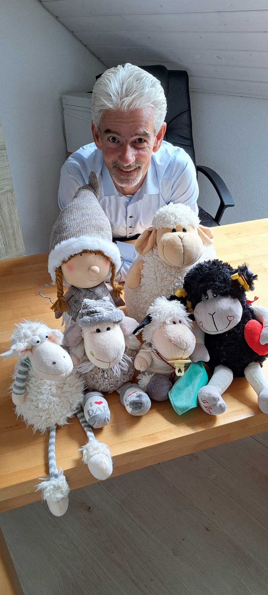 Foto von Autor Gerry Dressler, der lächelnd hinter einem Holztisch sitzt, auf dem die real existierenden Plüschfiguren seiner Buchcharaktere aus dem Winnieversum versammelt sind. Zu sehen sind das Schaf Winnie mit Schlafmütze und „I ♥ DAD“-Socken, das Brillenschaf Möbi, das schwarze Schaf Hilde mit rotem Filzherz, das Babyschaf Pfüddi mit Schnuller und das Grinseschaf mit erhobenem Huf sowie die kleine Schnürpelfee mit Zöpfen und Mütze.